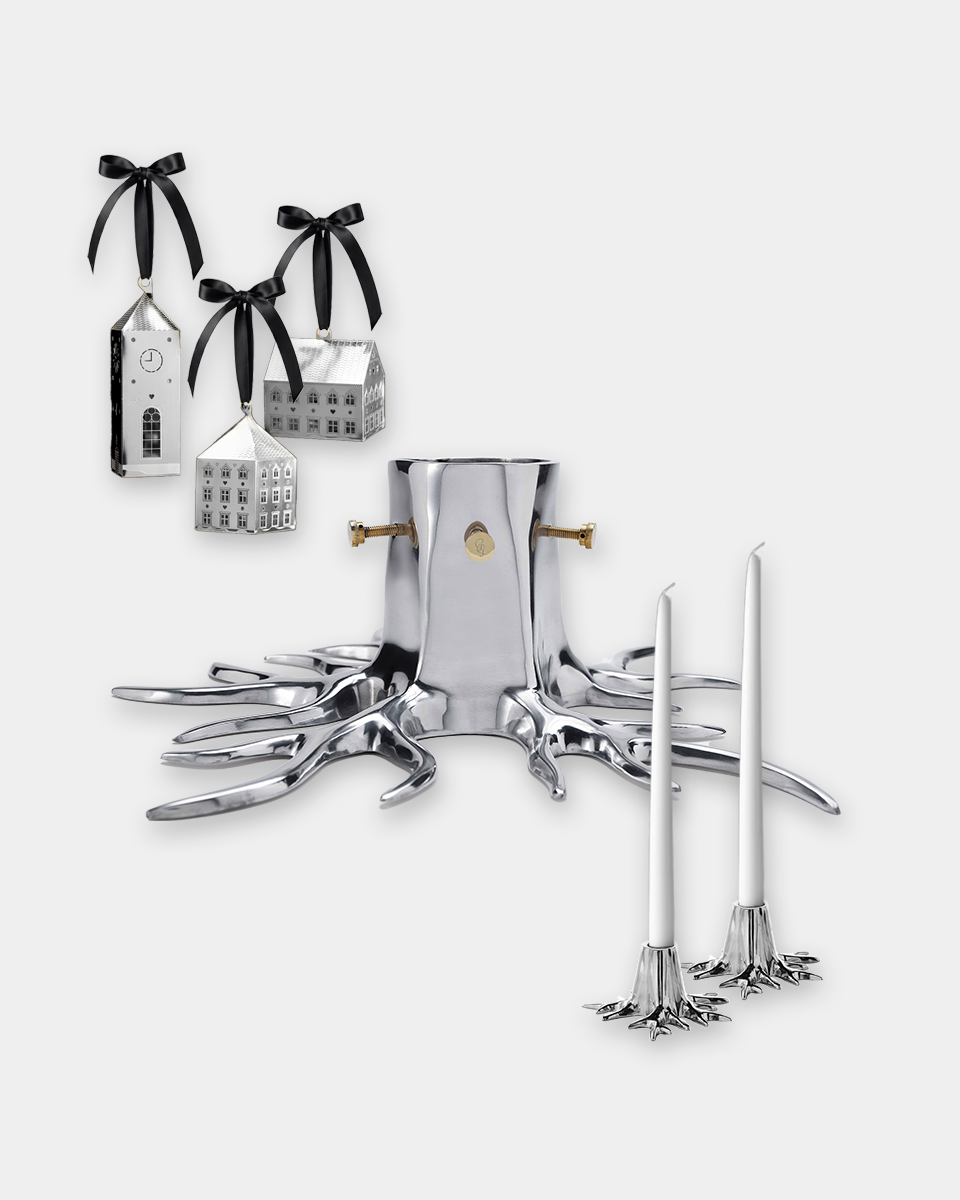 A silver metal holiday tree ornament set on a white background, including a tree stump base with antler-like feet, two taper candles, and three small hanging ornament houses tied with black ribbons.