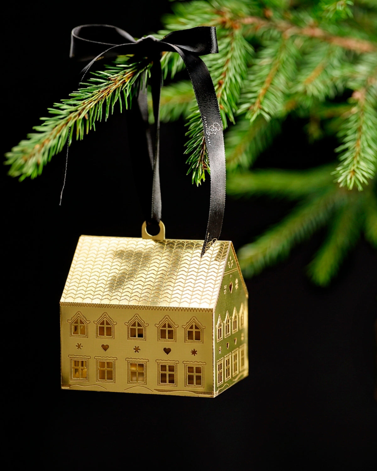 A gold-toned metal ornament shaped like a house, hanging from a black ribbon bow against a dark background with green pine branches above.