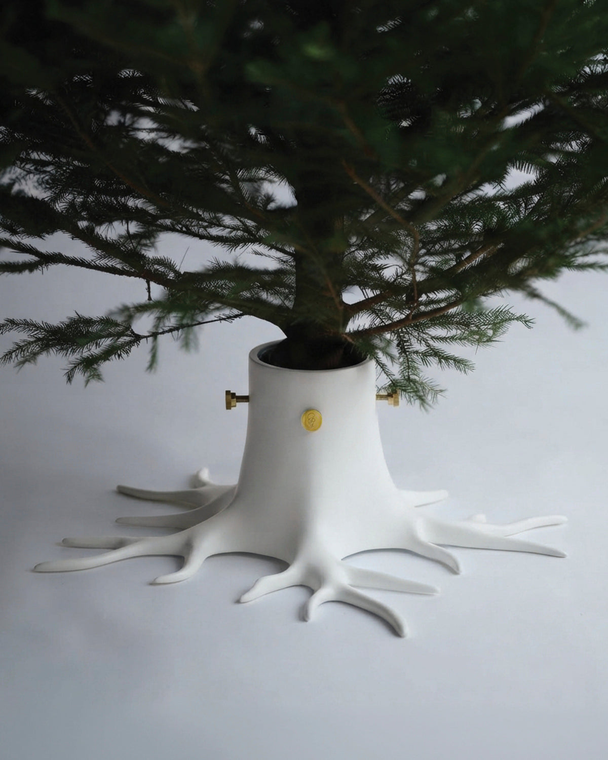 A white tree-shaped tree stand or planter with root-like legs and a cylindrical center, holding the base of a green Christmas tree. Small gold metal accents are visible on the stand against a plain light gray background.