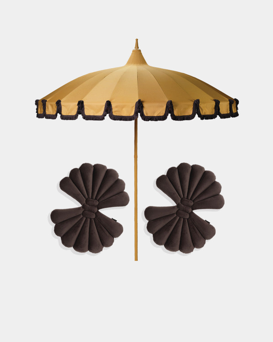 A saffron parasol-style umbrella with brown fringe trim and a saffron pole, shown above a pair of dark brown shell cushions accessories on a light gray background.