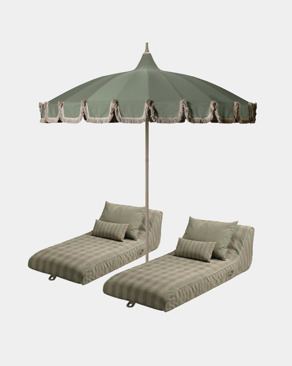 An outdoor patio set featuring a large green parasol with fringe trim, positioned behind two matching striped sun loungeers with pillows, set against a solid light gray background.