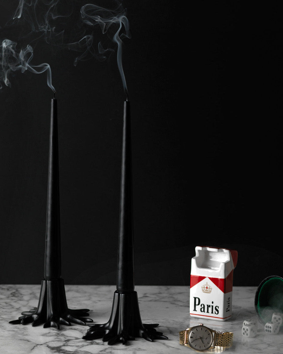 Two black candles with smoke, a box of Paris cigarettes, and a watch on a marble surface.