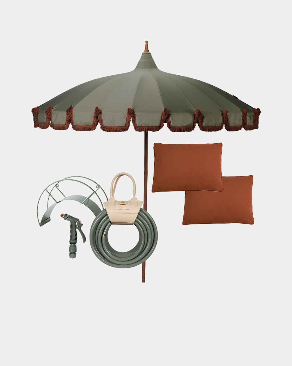 Outdoor patio umbrella set with a large olive green canopy featuring rust-colored fringe, a matching green garden hose, a metal sprinkler attachment, two rust-orange throw pillows, and a beige handbag, all arranged on a light gray background.