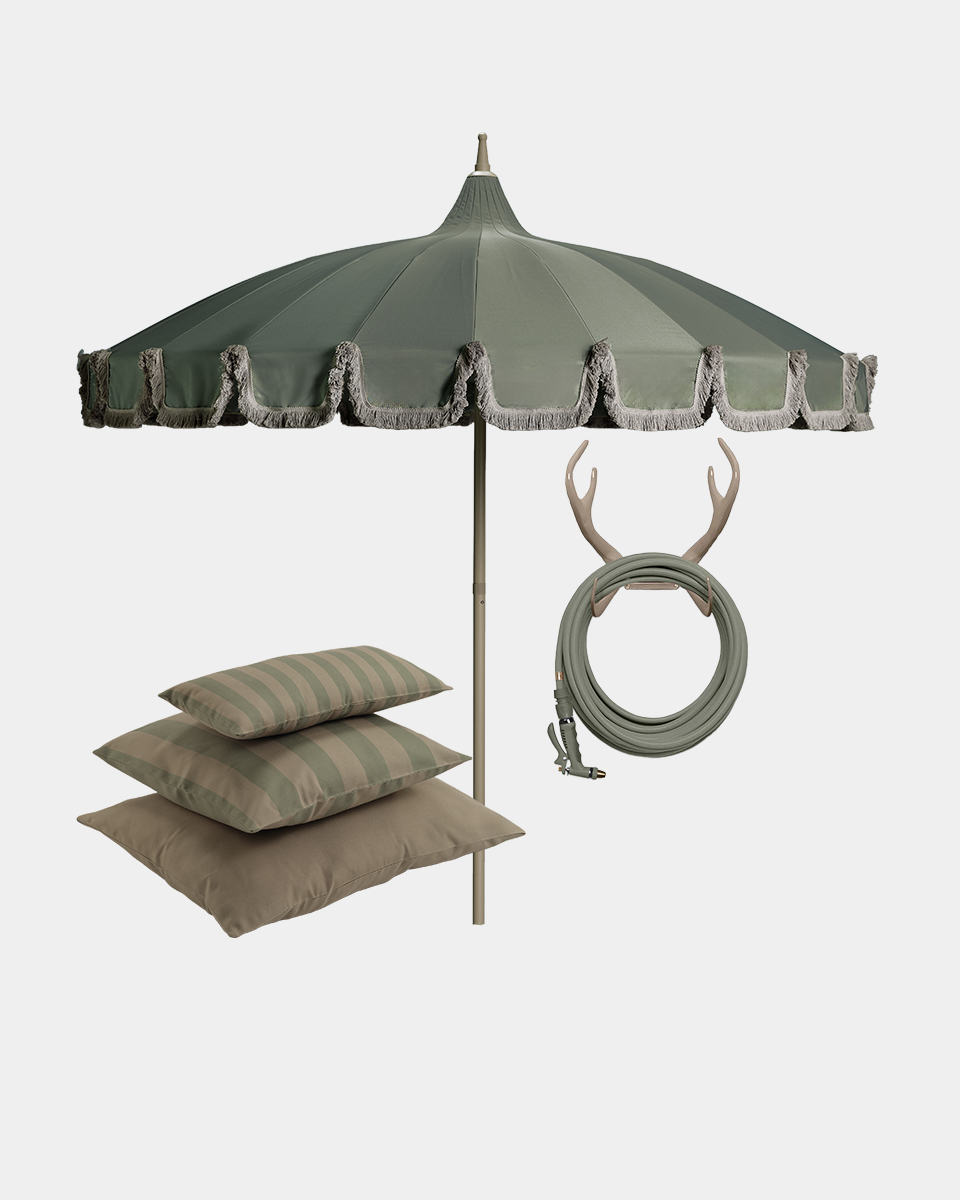 A large patio umbrella in muted sage green with fringe trim, shown with three coordinating pillows in beige and green stripes, a coiled green hose, and a pair of antler wall hooks against a plain light gray background.
