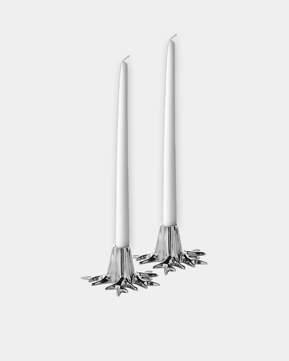 Two white taper candles in ornate silver metal bases with a starburst or snowflake-like bottom design, shown on a light gray background.