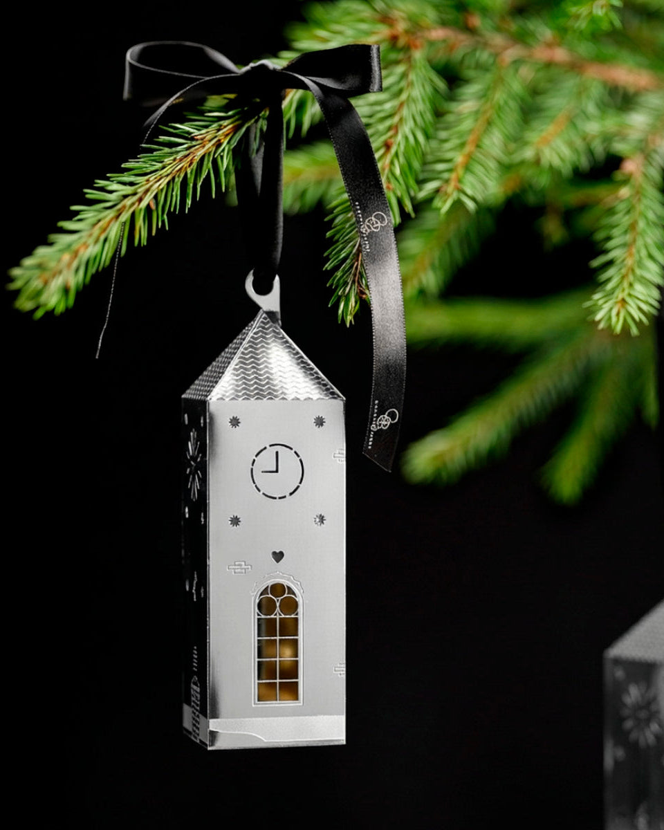 A silver metal Christmas ornament shaped like a church tower, hanging from a black ribbon bow against a dark background with blurred green pine branches.