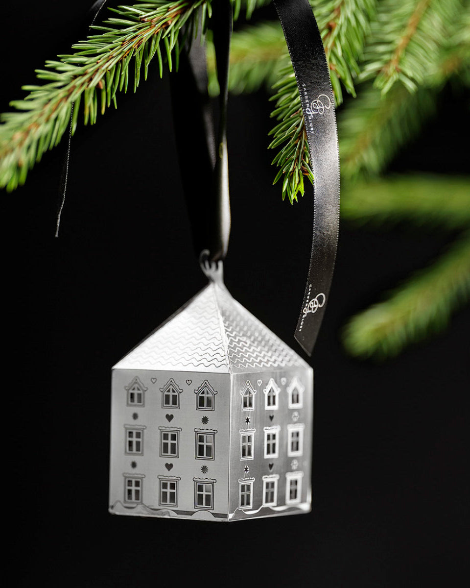 A silver house-shaped Christmas ornament with detailed window cutouts, hanging from a black ribbon under pine tree branches against a black background.