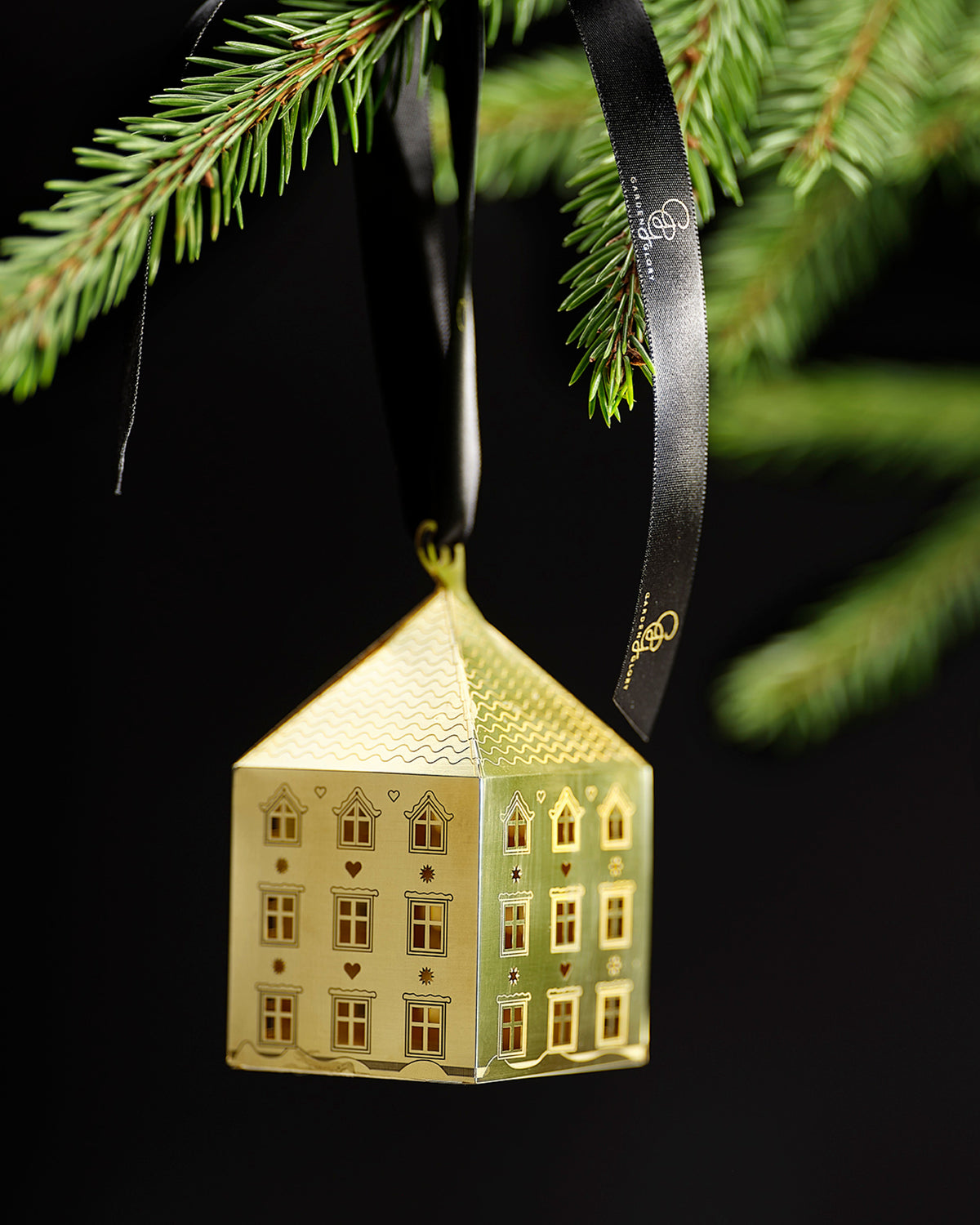 A gold-colored Christmas ornament shaped like a house, hanging from a black ribbon and displayed on a green pine tree branch against a black background.