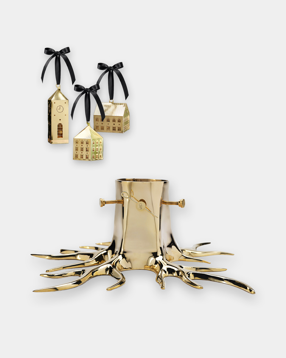 A gold-toned metal Christmas tree stump candle holder with root-like branches, shown on a light gray background. Two smaller hanging ornaments shaped like houses with black ribbons are displayed above it.